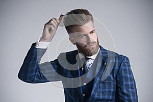 Confident in his perfect style. Confident young bearded man combing his hair and looking at away while standing against grey