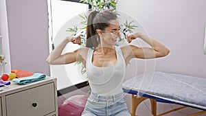 Confident hispanic woman flaunting power through strong arms gesture, smiling beautifully in young joy at rehab clinic