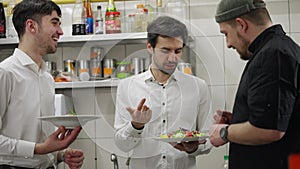 Confident and joyful waiters communicate with the chef about the salads he prepared in the kitchen in a restaurant. Guys