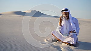 Confident male Arabian UAE Sheikh builder speaks on cellular with customer, sitting on sand with paper drawing in large