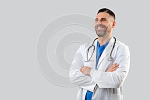 Confident male doctor in uniform posing with folded arms over grey studio background, looking at free space