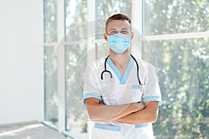 Confident male doctor wearing medical mask with arms crossed in hospital background