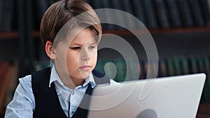 Confident male kid pupil chatting typing e learning use laptop library bookshelves interior closeup
