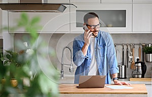 Confident male professional talking over smart phone while using laptop on kitchen counter at home