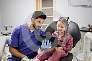 Confident male smiling professional dentist showing tablet pc computer to happy kid patient