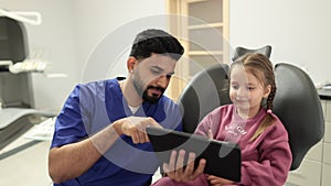 Confident male smiling professional dentist showing tablet pc computer to happy kid patient