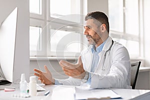 Confident man doctor working on computer at clinic, looking at screen, gesturing, having video conference with patient