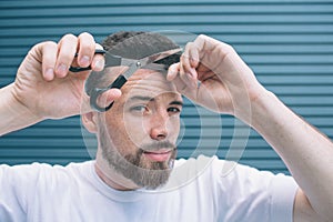 Confident man is standing and holding part of hair in one hand and scissors in another one. He is going to cut off bang