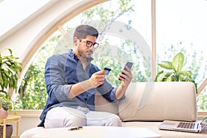 Confident man using mobile phone and credit card at home and paying bills online