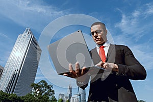 Confident man walking outdoors with laptop wearing black suit, glasses and red tie.