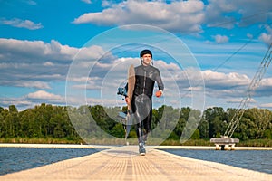 Confident man in wetsuit with wakeboard walking on floating bridge after training