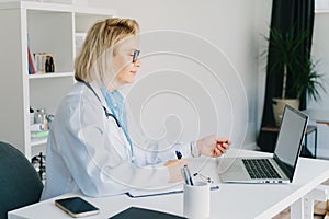 Confident mature caucasian senior female doctor in white coat using laptop for telemedicine with patients at hospital