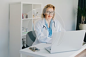 Confident mature caucasian senior female doctor in white coat using laptop for telemedicine with patients at hospital