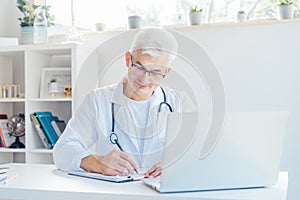 Confident mature caucasian senior male doctor making notes while using laptop for telemedicine with patients at hospital
