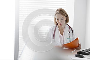 Confident mature doctor reading file at desk in hospital