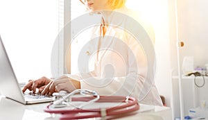 Confident mature doctor using laptop at table in hospital with yellow lens flare in background