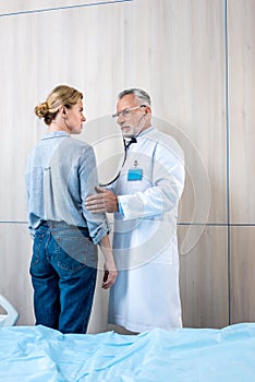 confident mature male doctor examining female patient by stethoscope