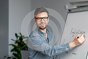 Confident mature male teacher giving online class, explaining new material, writing English rules on blackboard