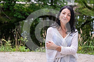 A confident mature woman in a light gray wrap smiles warmly, standing against a lush park setting, exuding self assured