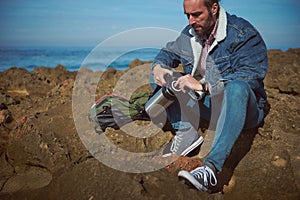 Confident mid adult bearded traveler man opening the stainless steel thermos flask, enjoying happy day alone by sea