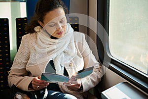 Confident mid adult multi ethnic woman enjoying train ride and reading a leaflet or book, sitting by train window