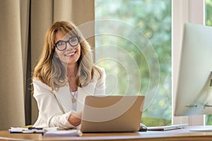 Confident mid aged business woman sitting at desk and using laptop for work photo