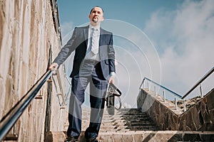 Confident middle age businessman with briefcase walking upstairs. Close-up of businessman wearing blue suit holding bag