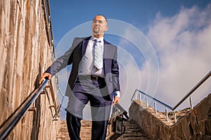 Confident middle age businessman with briefcase walking upstairs. Close-up of businessman wearing blue suit holding bag