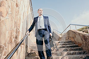 Confident middle age businessman with briefcase walking upstairs. Close-up of businessman wearing blue suit holding bag