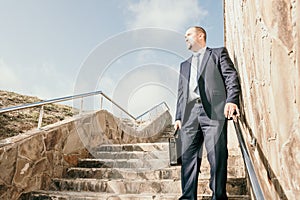 Confident middle age businessman with briefcase walking upstairs. Close-up of businessman wearing blue suit holding bag