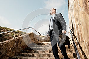 Confident middle age businessman with briefcase walking upstairs. Close-up of businessman wearing blue suit holding bag