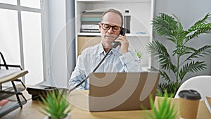 Confident middle age man animatedly talking on phone in office, handling business work on laptop with smiling gusto