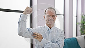 Confident middle age man flashing a handsome, healthy smile while making a strong muscle gesture in his cozy home living room