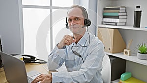 Confident middle age man, a smiling business worker, using laptop and headphones working on his job at the office, managing online