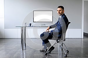 Confident middle aged businessman in suit sitting on desk with computer monitor with blank screen at office, mockup