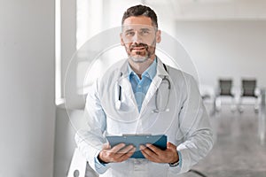 Confident middle aged male doctor in uniform holding clipboard and looking at camera, therapist man standing in clinic