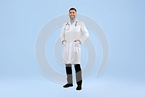 Confident middle aged male doctor in white coat posing with hands in pockets on blue background, full length