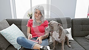 Confident middle-aged woman, with her grey-haired dog, enjoys relaxing on the sofa reading literature, expressing joy and comfort