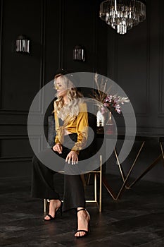 Confident model girl with long blond hair wearing fashionable outfit poses in dark luxurious interior