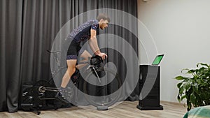 Confident motivated male in bicycle apparel cycling out of saddle on home smart bike trainer. Man doing cycling exercises and prep