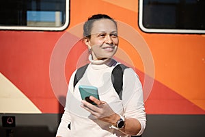Confident multiracial pretty woman, smiling looking aside, holding mobile phone in hand, standing at a railway station.