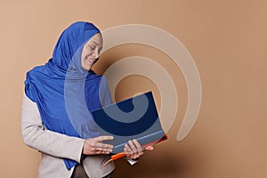 Confident Muslim woman in hijab, smiling, holding a pile of hardcover books in her hands, isolated on beige background