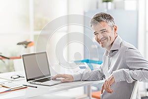 Confident office worker sitting at office desk and working with a laptop, he is smiling at camera