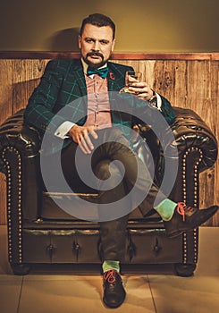 Confident old-fashioned man sitting in comfortable leather chair with glass of whisky in wooden interior at Barber shop.