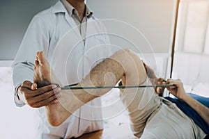 Confident physical therapist helps patient use resistance band stretching out his leg in clinic room