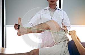 Confident physical therapist helps patient use resistance band stretching out his leg in clinic room