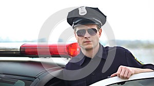 Confident police officer in uniform and sunglasses standing near patrol car