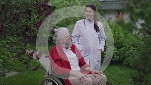 Confident positive Caucasian young woman talking with handicapped old man in wheelchair standing in garden outdoors