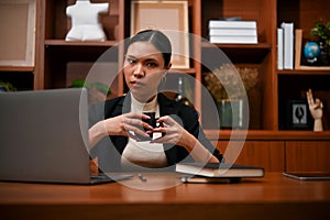 Confident and professional Asian businesswoman holding a coffee cup, sitting at her office desk