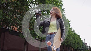 Confident redhead Caucasian retro woman walking with skateboard filming on vintage camera smiling. Tracking shot